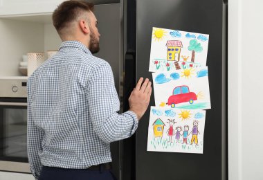Mutfakta çocuk resimleri olan buzdolabının kapısını açan adam.