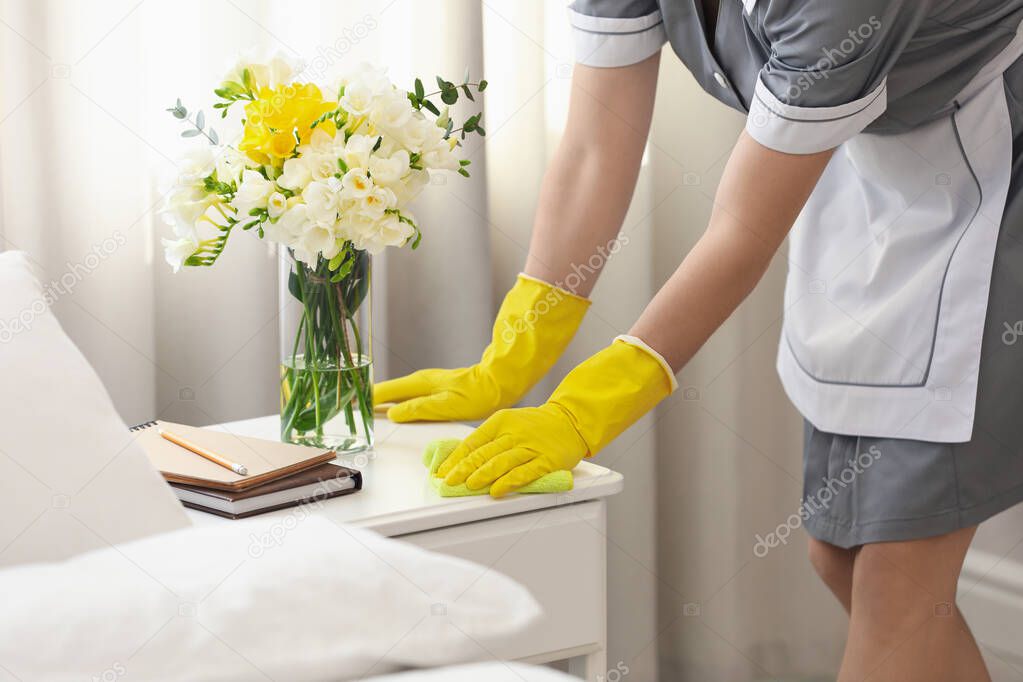 Young chambermaid wiping dust from nightstand in bedroom, closeup