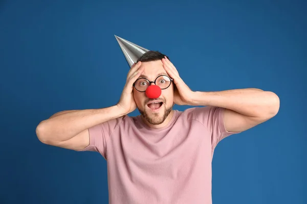 青い背景に眼鏡 パーティー帽子 ピエロの鼻を持つ感情的な男 4月のバカの日 — ストック写真