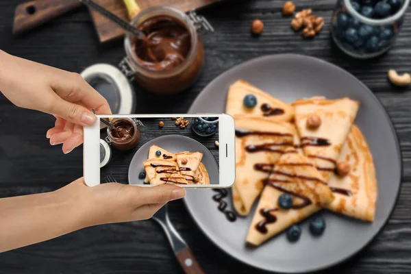 Food blogger taking picture of delicious thin pancakes at wooden table, closeup