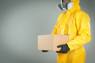 Kimyasal koruyucu giysili bir adam açık gri arka planda karton bir kutu tutuyor, yakın plan görüntüsü ve mesaj için yeri var. Virüsün yayılmasını önlemek