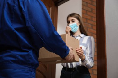 Tıbbi maske takan genç bir kadın içeride teslimatçıdan paket alıyor. Virüsün yayılmasını önlemek