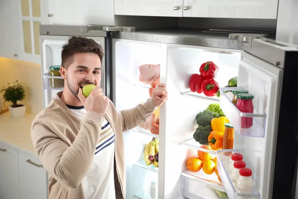 Mladý Muž Jíst Jablko Blízkosti Otevřené Ledničky Kuchyni — Stock fotografie