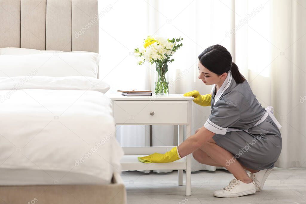 Young chambermaid wiping dust from nightstand in bedroom