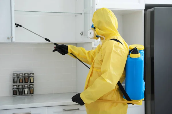 Pest control worker in protective suit spraying insecticide on furniture indoors