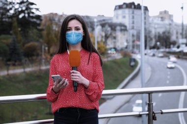 Sağlık maskesi, mikrofonu ve akıllı telefonu olan genç bir gazeteci şehir caddesinde çalışıyor. Virüs koruması