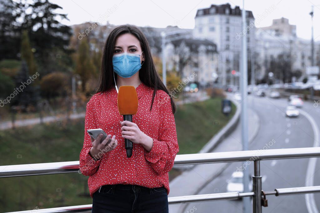 Young journalist with medical mask, microphone and smartphone working on city street. Virus protection