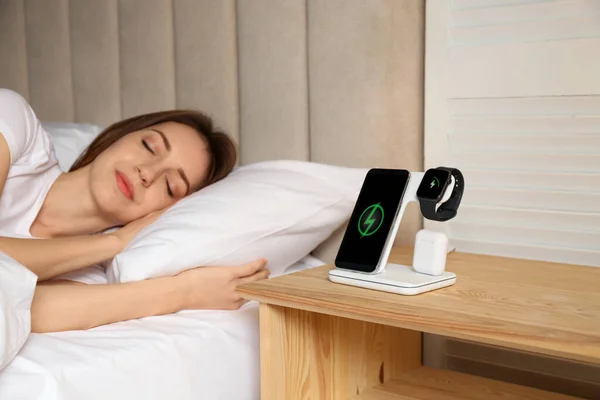 Smartphone, watch, earphones charging on wireless pad and sleeping woman in room