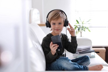 Kulaklıklı ve akıllı telefonlu şirin çocuk evde sesli kitap dinliyor.