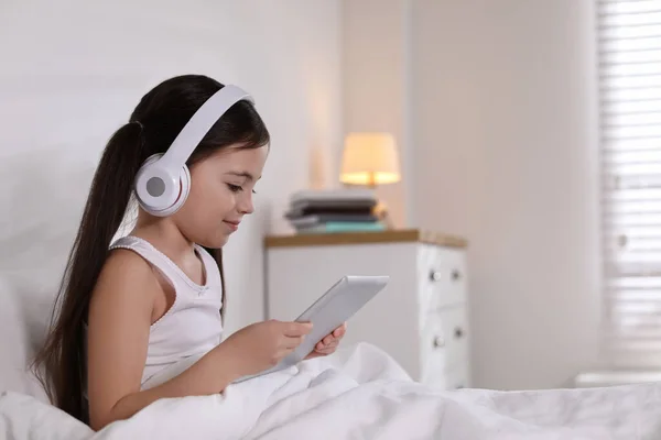 可爱的小女孩 带着耳机和平板电脑 在床上听有声读物 — 图库照片