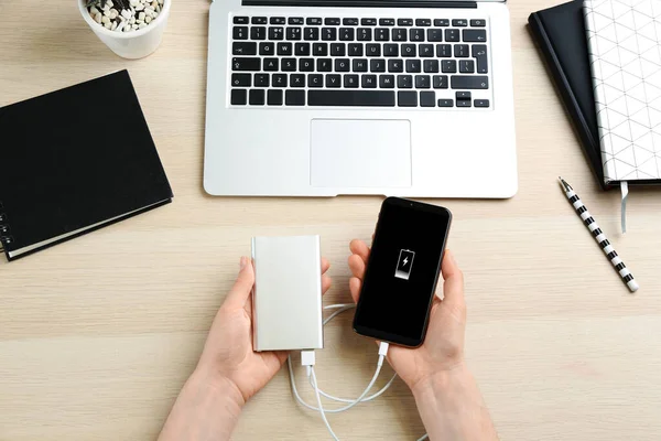 Woman charging mobile phone with power bank at wooden table, top view