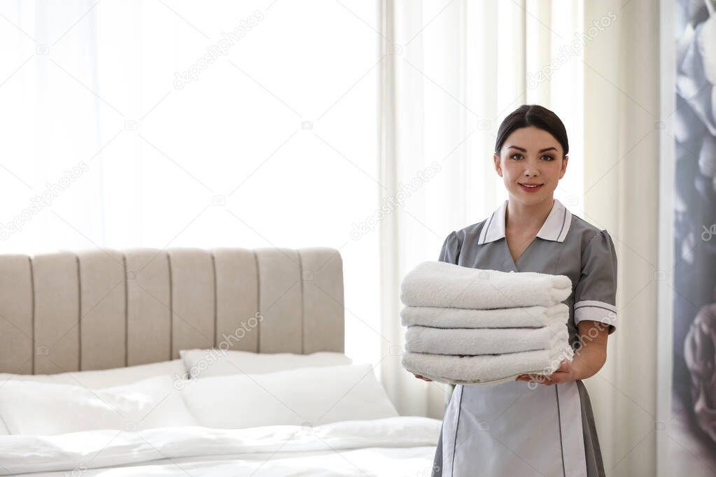 Young chambermaid holding stack of fresh towels in bedroom. Space for text
