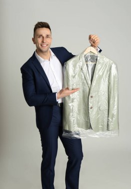 Man holding hanger with jacket in plastic bag on light grey background. Dry-cleaning service