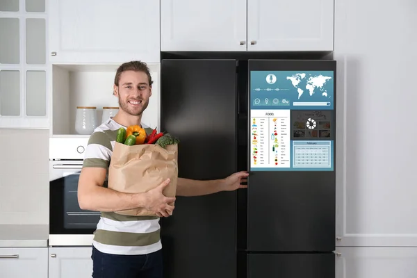 Young man with bag of groceries near smart refrigerator in kitchen