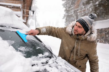 Genç adam kış günü arabanın ön camından kar temizliyor.
