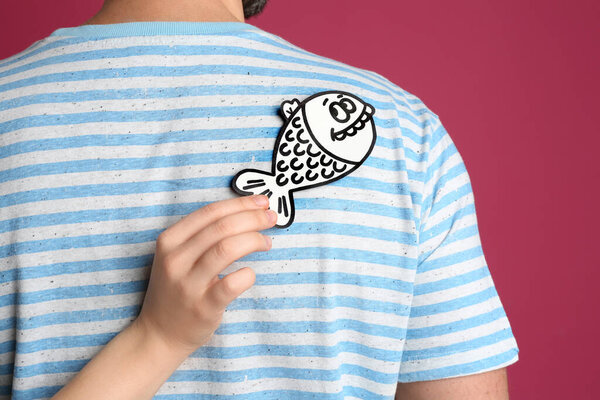 Woman sticking paper fish to friend's back on pink background. April fool's day