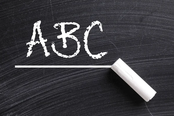 Piece of white chalk and letters ABC written on blackboard, top view