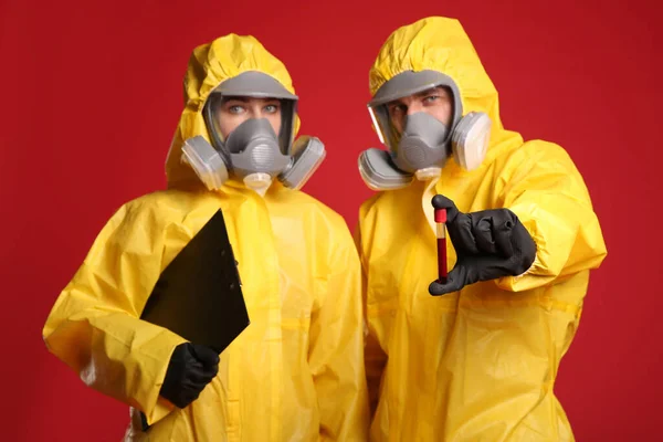 血液サンプルと赤の背景にクリップボードと化学保護スーツの人々 ウイルス研究 — ストック写真