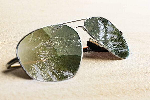 Stylish sunglasses with reflection of palm trees on white sand, closeup 