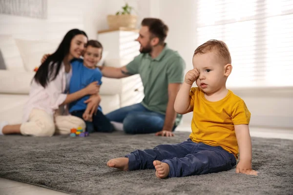 Unhappy baby sitting alone on floor while parents spending time with his elder brother at home. Jealousy in family