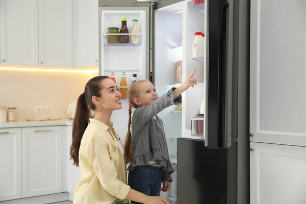 年轻的母亲带着女儿在厨房开着的冰箱旁 — 图库照片