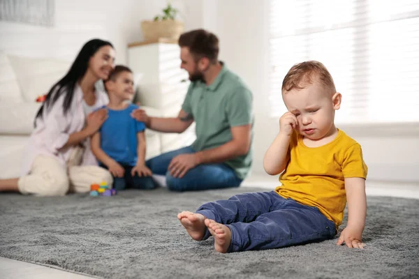 Unhappy baby sitting alone on floor while parents spending time with his elder brother at home. Jealousy in family