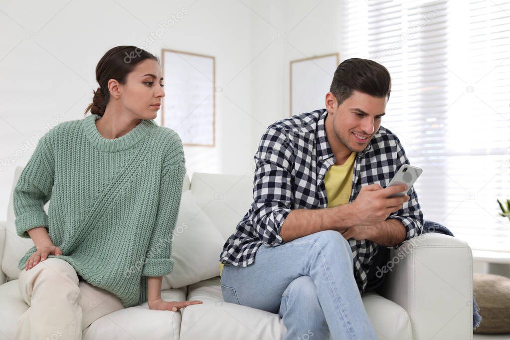 Distrustful woman peering into boyfriend's smartphone at home. Jealousy in relationship