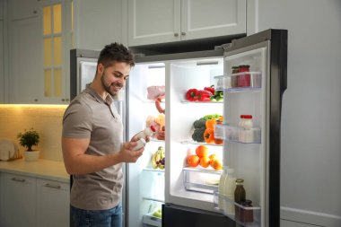 Mutfaktaki açık buzdolabının yanında yoğurt şişesi olan genç bir adam.