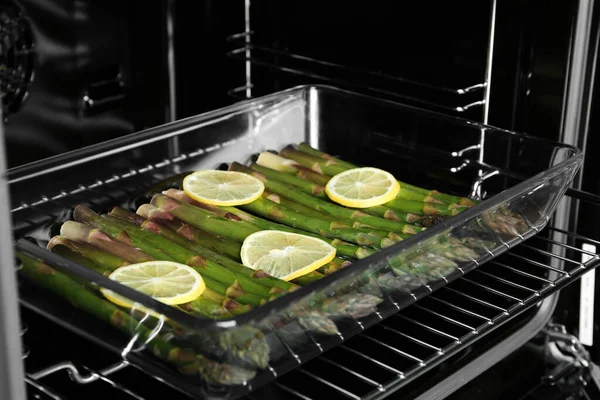 Raw asparagus with lemon slices in glass baking dish on oven rack, closeup