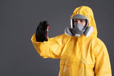 Kimyasal koruyucu elbiseli bir kadın gri arka planda test tüpü kan örneği tutuyor. Virüs araştırması