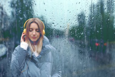 Kulaklıklı güzel bir kadın yağmurlu bir günde şehir caddesinde müzik dinliyor, ıslak pencereden bakıyor.