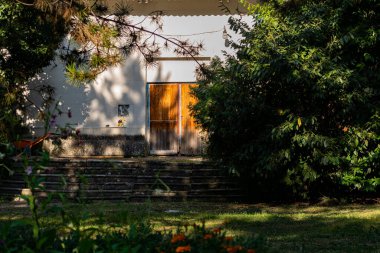 Parlak kahverengi, kırmızı ahşap kapılar, binanın girişi, çalıların ve yeşil ağaçların arasında geniş kapalı kapılar. Girişin açılışına giden basamakları tırmanıyorum.