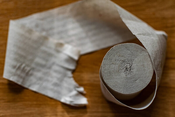 A roll of clean, plain toilet paper, cheap quality, dirty color, lies on a brown wooden table, slightly unwound and with the end torn off