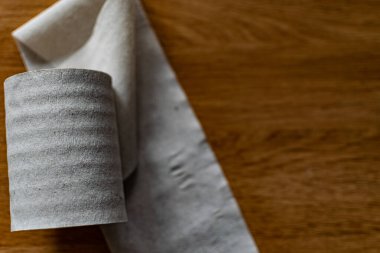 Bir rulo temiz, sade tuvalet kağıdı, ucuz kalite, kirli renk, kahverengi ahşap bir masada yatıyor, hafifçe yaralanmamış ve ucu koparılmış.