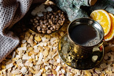 Yüzeyinde baloncuklar olan koyu kahverengi kahve tabakta, tam tahıllı, soyulmuş ve kurutulmuş portakalların yanında. Kızılağaç çiplerinin arkaplanı ve arka planda ağaç kabuğuyla ahşap çıkıntıları