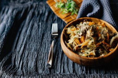 İstiridye mantarlı lahana, havuç ve soğanlı domuz dili. Tahta bir standın üzerinde, bir çatalın, bir dal çimenin ve bir peçetenin yanında, ahşap bir tabakta sıcak bir tabak. Rahatlamalı siyah arkaplan