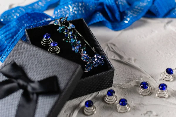 Beautiful brooch with blue and aquamarine stones and rhinestones of different sizes, metal base pins in a gift box with a black bow, around the ribbon with silver threads and springs.