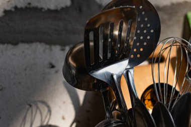 Beyaz bir stantta metal çatal bıçak takımı. Gümüş kaşıklar, çatallar ve paslanmaz çelik spatula mutfakta duruyor, arkasında da tahta ve rende var. Beton gri duvarlar çimentoyla.