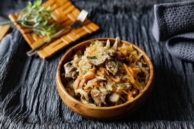 İstiridye mantarlı lahana, havuç ve soğanlı domuz dili. Tahta bir standın üzerinde, bir çatalın, bir dal çimenin ve bir peçetenin yanında, ahşap bir tabakta sıcak bir tabak. Rahatlamalı siyah arkaplan