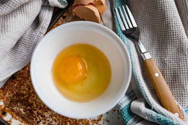 Çiğ yumurta yumurta yumurta, yumurta sarısı ve protein ile beyaz bir kaseye, kabuğun yanında, paslı metal bir arka planda çatal ve havlu, köyde olduğu gibi, basit ve sıradan her şey