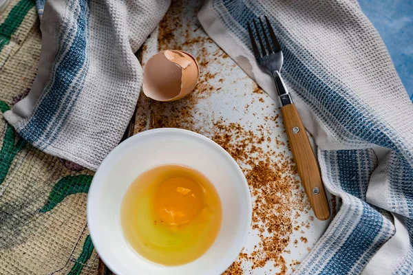 Raw egg broken into a white bowl with yolk and protein, next to the shell, fork and towel on a rusty metal background as at home in the village, everything simple and ordinary