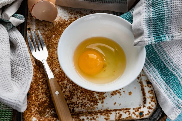 Raw egg broken into a white bowl with yolk and protein, next to the shell, fork and towel on a rusty metal background as at home in the village, everything simple and ordinary