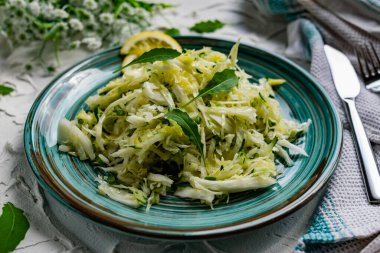 Taze sebzeli çıtır salata. Limonla süslenmiş salatalık ve roka yapraklı lahana ve çatal bıçaklı, hafif desenli taş bir masada bir buket beyaz çiçek.
