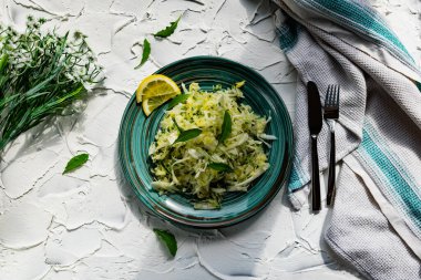 Taze sebzeli çıtır salata. Limonla süslenmiş salatalık ve roka yapraklı lahana ve çatal bıçaklı, hafif desenli taş bir masada bir buket beyaz çiçek.