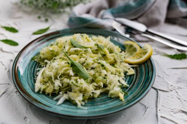 Taze sebzeli çıtır salata. Limonla süslenmiş salatalık ve roka yapraklı lahana ve çatal bıçaklı, hafif desenli taş bir masada bir buket beyaz çiçek.