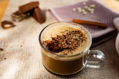 Köpüğünün üzerinde çikolata parçaları olan sıcak kapuçino, tarçın çubuğu, altın şeker boncukları ve yanında tatlı bir waffle. Şeffaf bir fincanda yumuşak bir bez üzerinde iç.