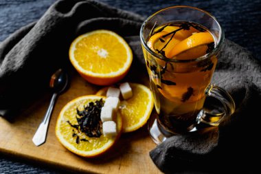 Citrus içeceği, şeffaf cam bardakta taçyapraklı çay, tatlı portakal dilimleri ve rafine şeker. Ahşap bir tahtada, yumuşak, koyu bir arka planda kahverengi bir peçeteyle meyve dilimlemek.