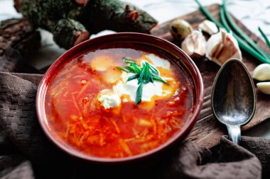 Kahverengi bir tabakta kırmızı borş, eski bir tahta tahtanın yanında sarımsak ve bıldırcın yumurtası, yeşil pırasa ve süzülen dallar. Çorbada beyaz ekşi krema, eski bir Polonya kaşığının yanında.