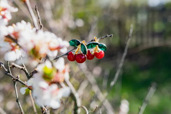 Cute little brooches in the form of red cherries with green leaves made of emerald green stones. In combination with different elements of nature, branches of a flowering Bush with white petals