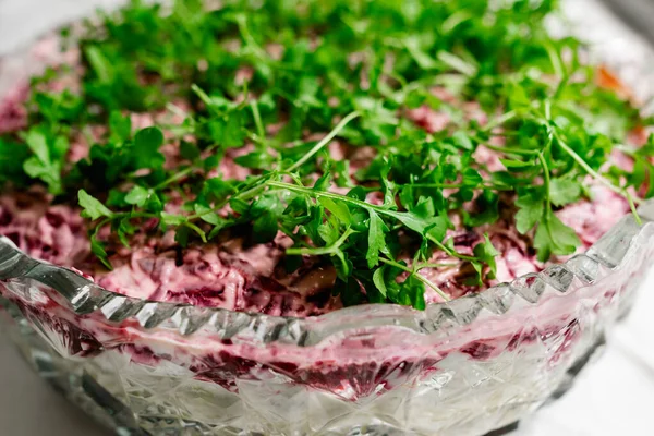 A large crystal cut-glass bowl with puff salad herring under a fur coat, a Russian dish with boiled beetroot and mayonnaise, green leaves of watercress on top. White textured stone background